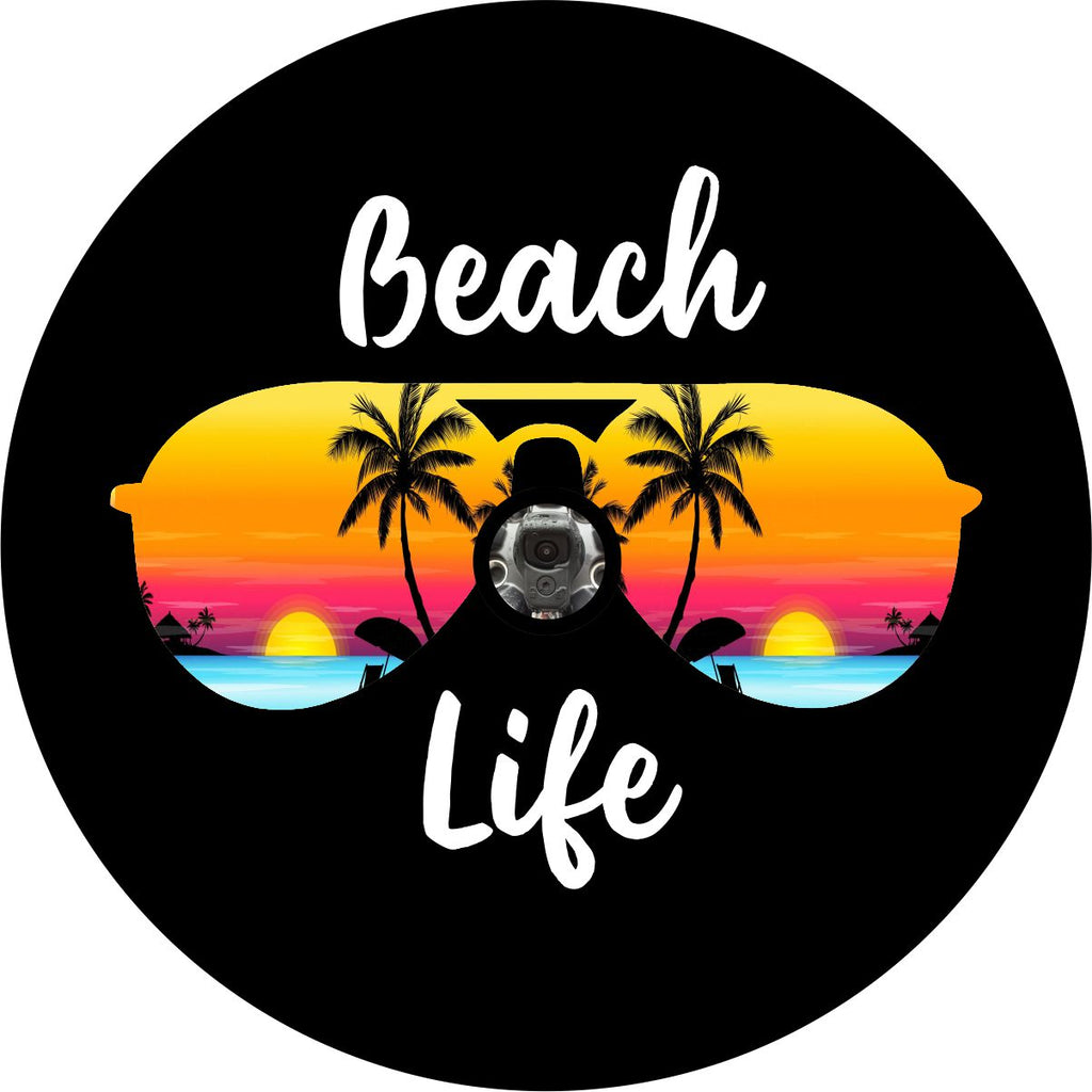 A unique spare tire cover for with the words beach life in cursive white font on black vinyl with the silhouette of aviator sunglasses and a tropical island beach sunset within the frames of the sunglasses. This beach scene spare tire cover is designed to accommodate a JL backup camera for Jeep Wrangler and other vehicles with backup cameras.