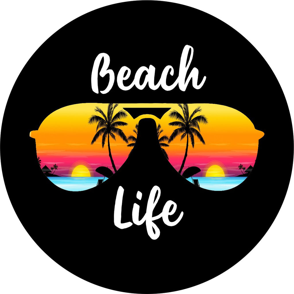 A unique spare tire cover with the words beach life in cursive white font on black vinyl with the silhouette of aviator sunglasses and a tropical island beach sunset within the frames of the sunglasses