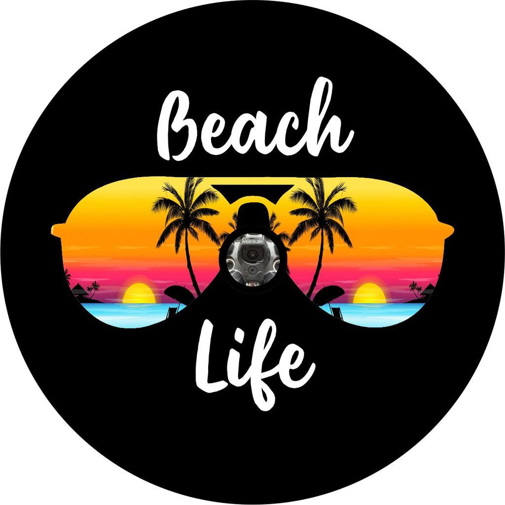 A unique spare tire cover for with the words beach life in cursive white font on black vinyl with the silhouette of aviator sunglasses and a tropical island beach sunset within the frames of the sunglasses. This beach scene spare tire cover is designed to accommodate a JL backup camera for Jeep Wrangler and other vehicles with backup cameras. 