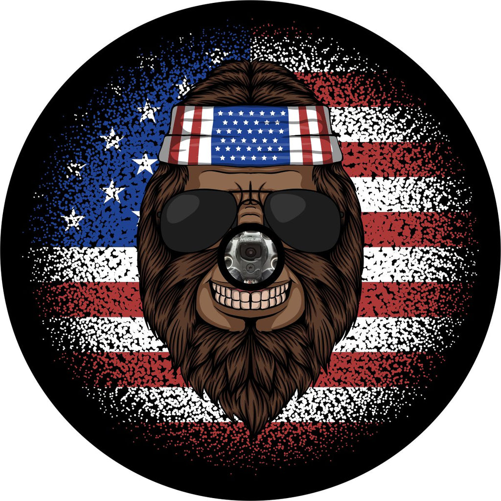 Smiling bigfoot sasquatch spare tire cover design. Bigfoot wearing an American flag bandana and sunglasses with the American flag is in the background with dispersion effect. Bronco, RV, camper, and Jeep tire cover with camera hole options available.